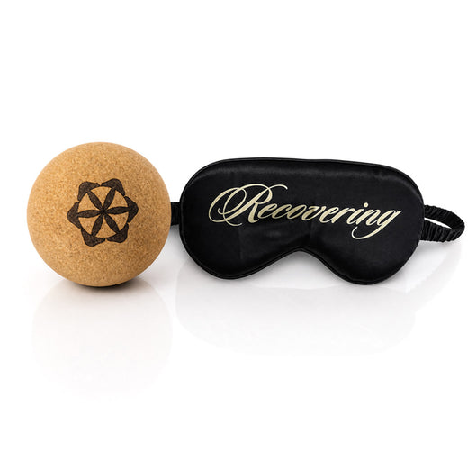 A cork lacrosse ball beside a black silk sleep mask reading Recovering